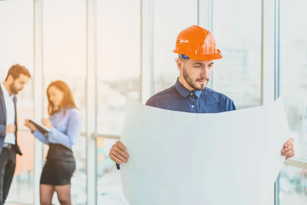 The architect is in a cask of orange color. In the office against the background of their colleagues.