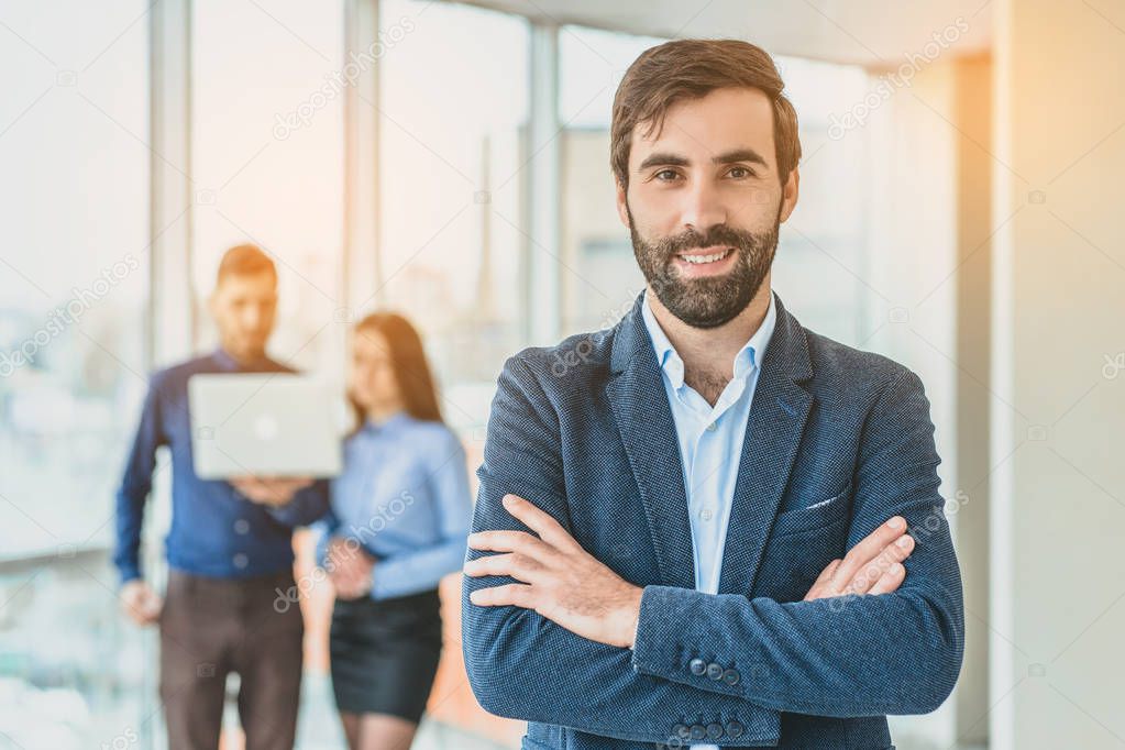 A businessman stands in the office folded his hand to his hand. During this time, he stands against the background of his colleagues who work in the laptop.