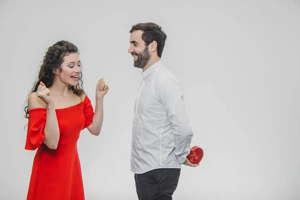 Casal jovem amando. Um homem fez um presente para a mulher. Durante este tempo, o campo vermelho é apresentado na forma de um coração. A esposa está agradavelmente surpreendida. Vestido com um vestido vermelho e uma camisa branca . — Fotografia de Stock