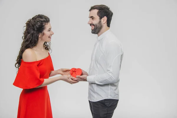 Casal jovem amando. Um homem fez um presente para a mulher. Durante este tempo, o campo vermelho é apresentado na forma de um coração. A esposa está agradavelmente surpreendida. Vestido com um vestido vermelho e uma camisa branca . — Fotografia de Stock