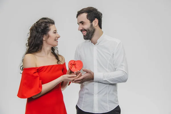 Jeune couple aimant. Un homme a fait un cadeau pour sa femme. Pendant ce temps, le champ rouge se présente sous la forme d'un cœur. La femme est agréablement surprise. Habillé d'une robe rouge et d'une chemise blanche . — Photo