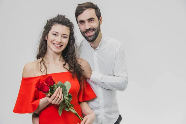 Homem elegante dá uma bela mulher um vestido vermelho e um buquê de rosas vermelhas. Casal amoroso em um fundo branco . — Fotografia de Stock