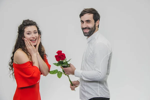 Um jovem escondendo um monte de rosas por trás de sua surpresa para surpreender sua namorada no Dia dos Namorados. Vestido com vestido vermelho e camisa branca . — Fotografia de Stock