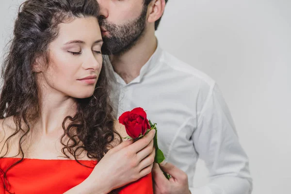 Un jeune et beau homme couvre doucement sa belle femme. Pendant ce temps, une rose rouge. La fille est vêtue d'une robe rouge, un gars en chemise blanche. Sur fond blanc . — Photo