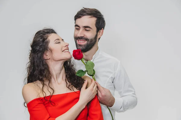 Um jovem e bonito homem gentilmente cobre sua bela esposa. Durante este tempo, uma rosa vermelha. A menina está vestida com um vestido vermelho, um cara com uma camisa branca. Em um fundo branco . — Fotografia de Stock