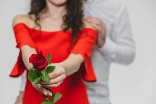 Un hombre joven y guapo cubre gentilmente a su hermosa esposa. Durante este tiempo, la esposa sostiene una rosa roja. La chica está vestida con un vestido rojo, un tipo con una camisa blanca . — Foto de Stock