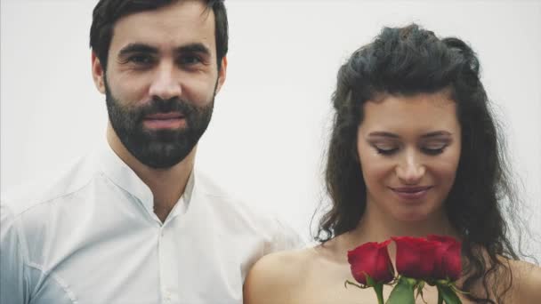 Hermoso chico elegante en una camisa clásica con rosas rojas en sus manos. Aparece sobre un fondo blanco. Le da las rosas a su guapa novia y la besa tiernamente. Día de San Valentín . — Vídeo de stock