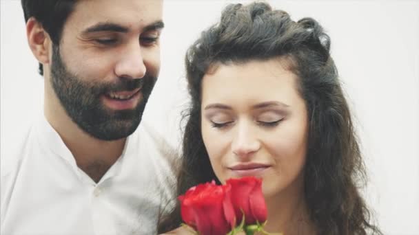 Hermoso chico elegante en una camisa clásica con rosas rojas en sus manos. Aparece sobre un fondo blanco. Le da las rosas a su guapa novia y la besa tiernamente. Día de San Valentín . — Vídeo de stock