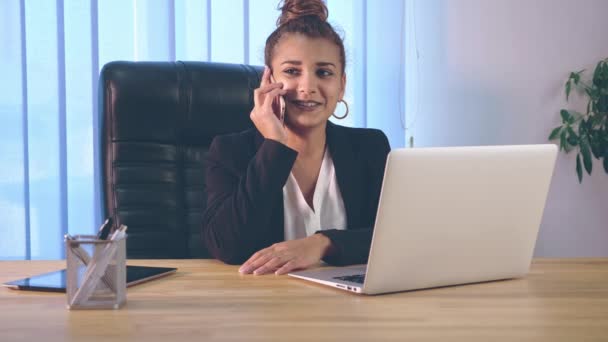 Dziewczyna jest ubrana w stylowe ubrania, siedział w biurze w pobliżu laptopa. W tym czasie mówi przez telefon. — Wideo stockowe