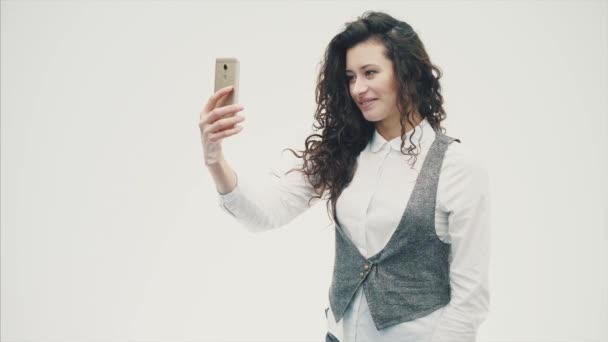 Una joven enérgica con el pelo rizado es fotografiada por teléfono. Habiendo hecho un sephi. Sobre un fondo blanco . — Vídeos de Stock