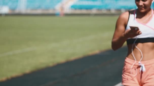 Retrato de una chica fuerte en ropa deportiva corriendo en el estadio. Hermosa chica corre en el estadio y escucha al jugador . — Vídeo de stock