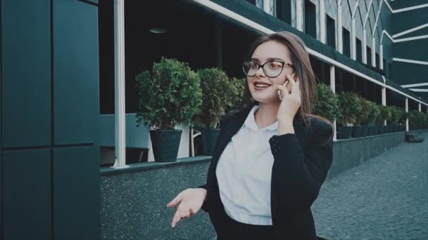 Νεαρή κοπέλα αρκετά έξυπνες επιχειρηματικές περπατώντας στα ρούχα επιχειρήσεων. Κατά τη διάρκεια αυτής μιλά στο τηλέφωνο. — Αρχείο Βίντεο