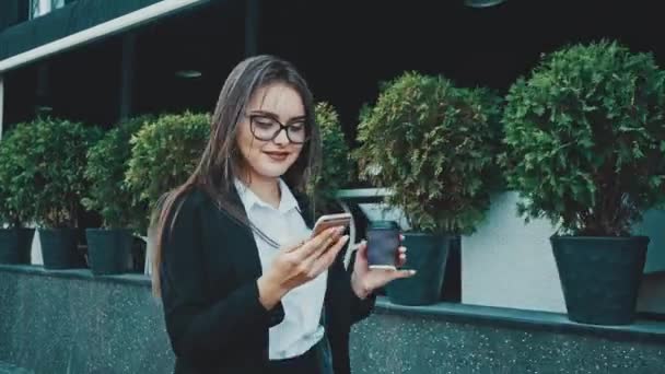 Jovem menina de negócios muito inteligente andando em roupas de negócios. Durante isso, ela mantém o café e olha para o telefone . — Vídeo de Stock