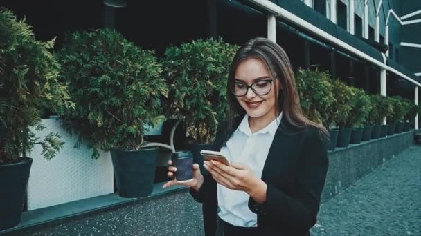 Giovane ragazza d'affari piuttosto intelligente che cammina in abiti da lavoro. Durante questo lei tiene il caffè e guarda nel telefono . — Video Stock