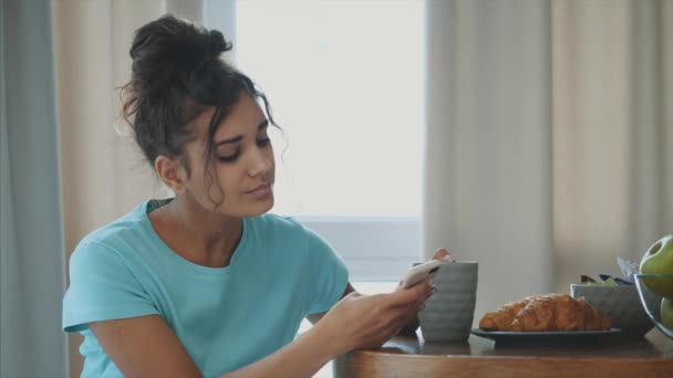 Jovem feliz mulher refrescante com xícara de café, lanches e frutas frescas na mesa da cozinha no início da manhã . — Vídeo de Stock