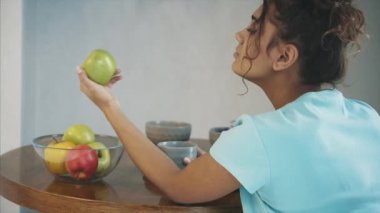 Bir elinde yeşil elma ve kruvasan sebze sağlıklı ve zararlı gıda arasında zor bir seçim yapmak mutfak masasının üzerinde arka plan üzerinde diğer elinde güzel genç ince esmer.