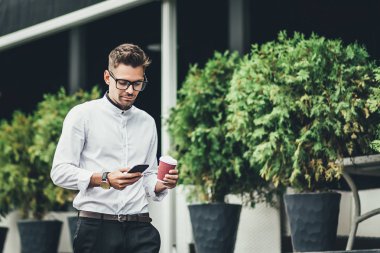 Bir kahve molası. Kahve fincanı tutarak ve açık havada yürürken akıllı telefonunu kullanarak bardaklarda kendine güvenen genç adam