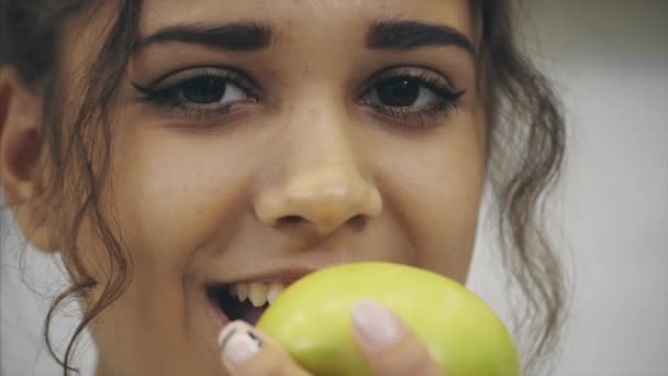 Beautiful young slender brunette with green apple in one hand and croissant in the other hand on background of vegetables on kitchen table making a difficult choice between healthy and harmful food. — Stock Video