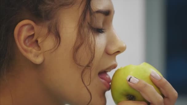 Beautiful young slender brunette with green apple in one hand and croissant in the other hand on background of vegetables on kitchen table making a difficult choice between healthy and harmful food. — Stock Video