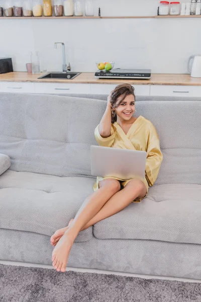 Brünette entspannt mit ihrem Laptop auf der Couch in ihrer Wohnung liegend. glückliche junge Frau, die es sich auf einem bequemen Sofa bequem macht und zu Hause einen Laptop benutzt. — Stockfoto