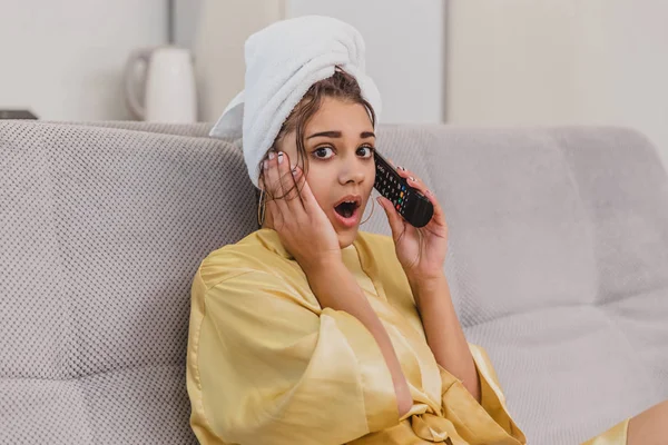 Eine glückliche Frau mit Fernbedienung, die einen Fernsehsender wählt, hebt die Fernbedienung zu den Ohren des Sprechens. während der Rest nach dem Bad. — Stockfoto