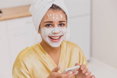 Spa uygulamaları evde yapıyor güzel ve genç birbayan. Telefonda onun elinde tutan bir maske ile oturur.