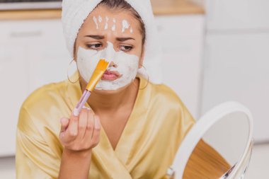 Yüz ve vücut cilt bakımı, yüz tedavi ev concept. Mutlu genç kadın bir fırçayla yüzünde beyaz bir maske uygulanıyor.