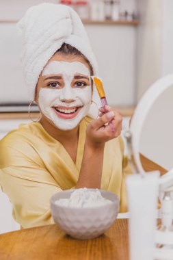 Yüz ve vücut cilt bakımı, yüz tedavi ev concept. Mutlu genç kadın bir fırçayla yüzünde beyaz bir maske uygulanıyor.