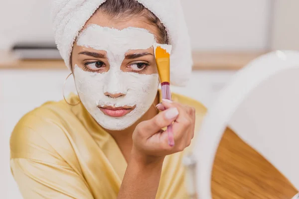 Face and body skin care, face treatment at home concept. Happy young woman applying a white mask on her face with a brush. — Stock Photo, Image