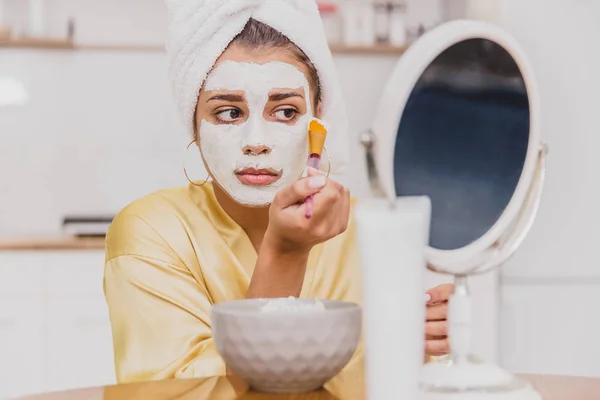 Soins de la peau du visage et du corps, traitement du visage à domicile concept. Joyeux jeune femme appliquant un masque blanc sur son visage avec un pinceau . — Photo