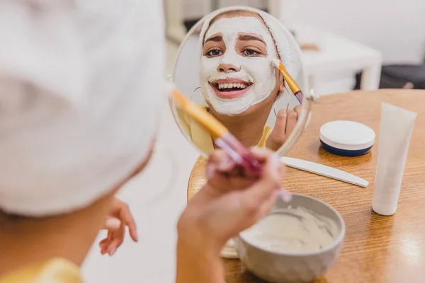 Wajah dan perawatan kulit tubuh, perawatan wajah di rumah konsep. Wanita muda yang bahagia mengenakan topeng putih di wajahnya dengan kuas . — Stok Foto