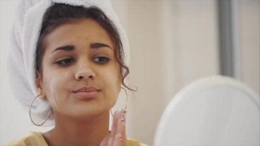 Genç kadın gülümsüyor. Uyanma spa sabah sahiptir. Mutlu kız başında bir havlu ile yatak odasında yüzünde bir maske. İyi cilt tonunu