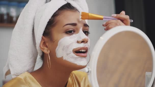 Portrait émotionnel d'une belle fille nue heureuse et calme avec un masque cosmétique en argile bleue sur la moitié de son visage, regardant le miroir de la caméra appliquer de l'argile avec un pinceau . — Video