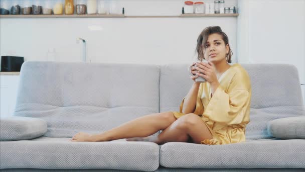 ソファの部屋でバスローブ姿の若い女性が座っています。彼の飲み物をコーヒー、ソファに座っているとテレビ居心地の良い家庭的な雰囲気を見て. — ストック動画