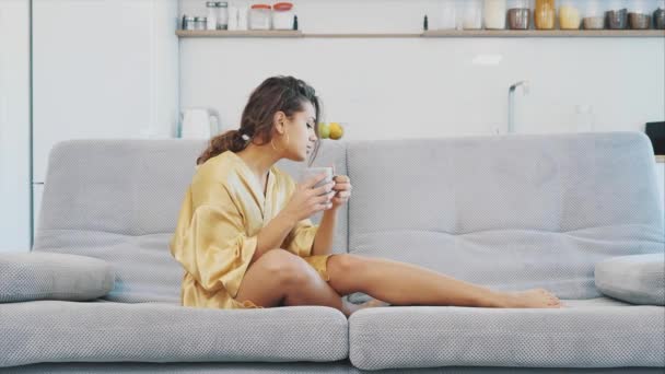 Mujer joven en un albornoz sentarse bien en la habitación en el sofá. Bebe café sentado en el sofá y viendo la tele. Ambiente acogedor en casa . — Vídeos de Stock