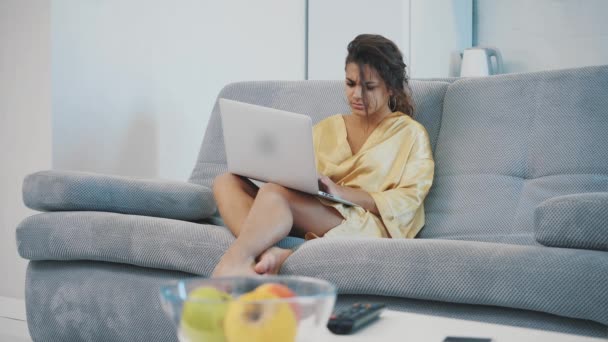 Mujer joven con una bata sentada en un sofá cama. Utiliza un ordenador portátil y está nervioso. Ambiente hogareño . — Vídeos de Stock