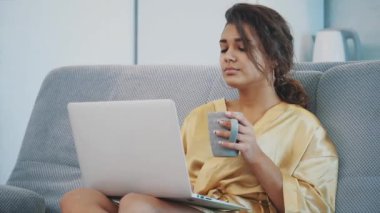 Esmer kanepede onun evinde yatarken onu laptop ile rahatlatıcı. Rahat bir kanepede rahatlatıcı ve evde bir dizüstü bilgisayar kullanarak mutlu genç kadın.
