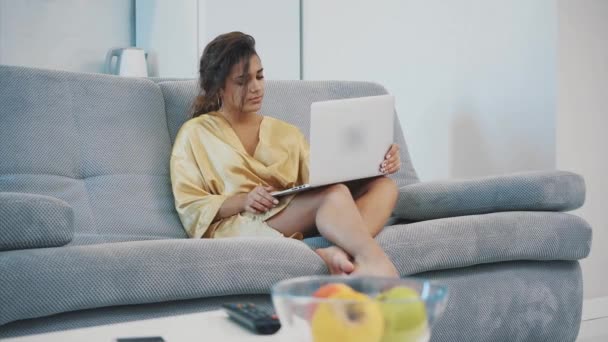 Brunette relaxando com seu laptop deitado no sofá em seu apartamento. Jovem feliz relaxando em um sofá confortável e usando um laptop em casa . — Vídeo de Stock