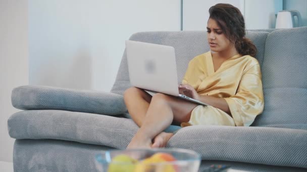 Morena relajándose con su portátil tumbada en el sofá de su apartamento. Mujer joven feliz relajarse en un cómodo sofá y el uso de un ordenador portátil en casa . — Vídeos de Stock