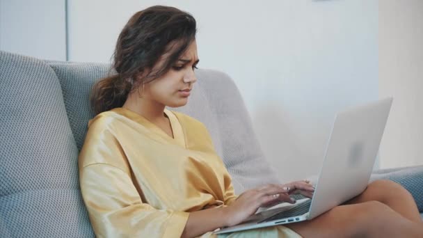 Brunett avkopplande med sin laptop liggande på soffan i hennes lägenhet. Glad ung kvinna koppla av på en bekväm soffa och använder en bärbar dator hemma. — Stockvideo