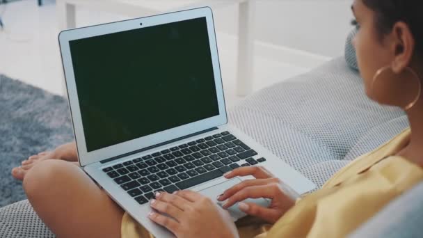 微笑的年轻女子在家里使用笔记本电脑, 她放松在舒适的沙发上。一个有吸引力的有趣的模型与电脑打字。绿色屏幕. — 图库视频影像