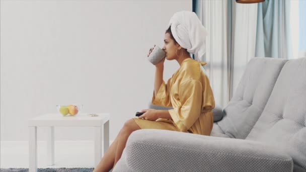 Giovane donna con una vestaglia e un asciugamano in testa. Si mette nella stanza sul divano e tiene tra le mani una cassetta nera e una tazza di caffè . — Video Stock