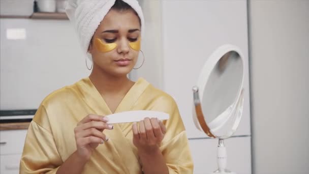 Jonge vrouw glimlacht. Wakker worden in de ochtend gelukkig meisje in een handdoek op haar hoofd met een masker op haar gezicht in de slaapkamer. Goede huidtoon. Perst nagelvijl. — Stockvideo