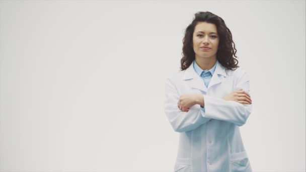 Porträt einer fröhlichen jungen lächelnden Ärztin. gekleidet in ein weißes Gewand. gleichmäßig stehend mit gekreuzten Händen auf grauem Hintergrund. — Stockvideo