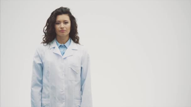 Porträtt av glada unga leende flicka läkare. Klädd i en vit klädnad. Jämnt stående med korsade händer på en grå bakgrund. — Stockvideo