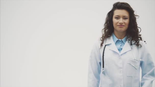 Ritratto di giovane donna felice sorridente medico. Vestita con una vestaglia bianca. Uniformemente in piedi con le mani incrociate su uno sfondo grigio . — Video Stock