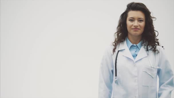 Retrato de feliz jovem sorridente médico menina. Vestido com um roupão branco. Evenly de pé com as mãos cruzadas em um fundo cinza . — Vídeo de Stock