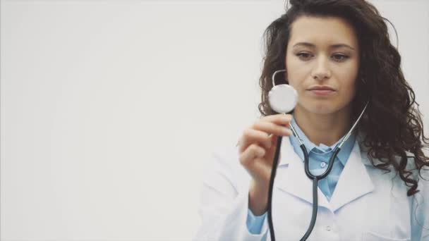 Portret szczęśliwy młody lekarz uśmiechający się dziewczyna. Ubrany w białą szatę. Równomiernie stojący na szarym tle. W tym czasie fonendoscope jest Szukam pilota poziomo. — Wideo stockowe
