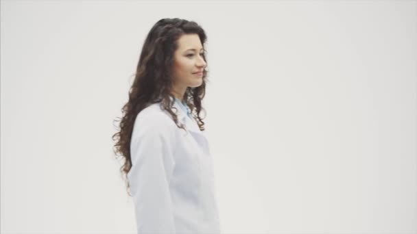 Portrait de heureuse jeune fille souriante médecin. Vêtue d'une robe blanche. Soirée debout sur un fond gris. Pendant ce temps, il regarde la caméra et montre un geste d'appel . — Video