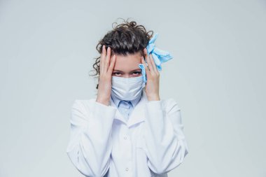 Genç bir kadın doktor maskeli giymiş. Aynı zamanda o kafasına eldiven ellerini tutar. Aynı zaman duygu ağrı. Sağlık ve tıp. Kavramları.